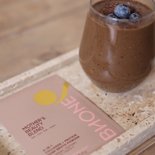 Glass of chocolate smoothie with blueberries on a stone surface, next to a box labeled 'Bone' and 'Mother's Beauty Blend'.