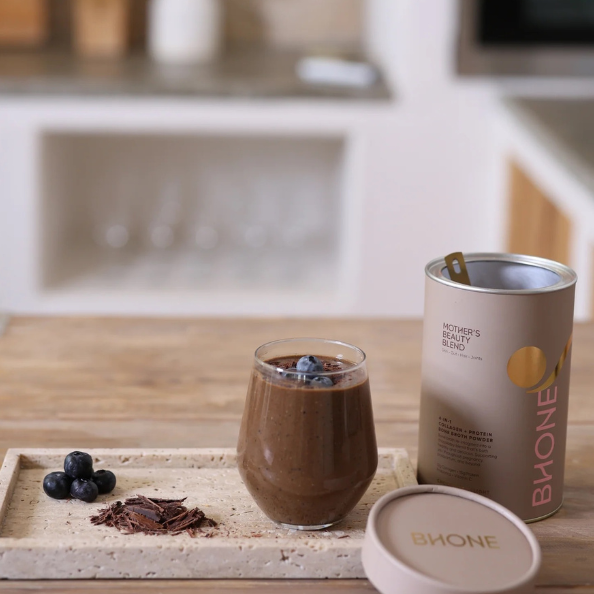 Glass of chocolate smoothie with blueberries, a can of Buone, and a container on a wooden surface.