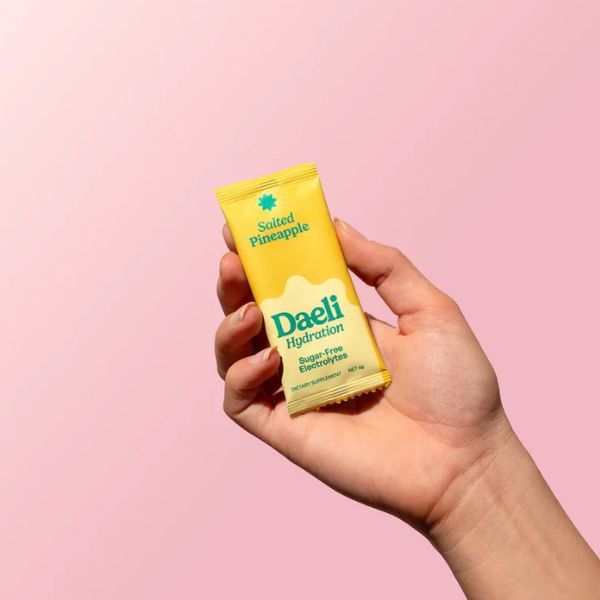 Hand holding a yellow Daeli Hydration packet against a pink background